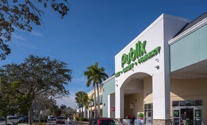 Publix supermarket at Freedom Square in Naples, FL, shown with a clean arched entrance, green signage, landscaped walkways, and shoppers moving through the parking area in front of the store.