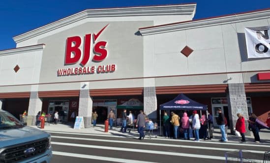 Exterior of a BJ’s Wholesale Club with shoppers gathered outside near pop-up tents under a clear blue sky.