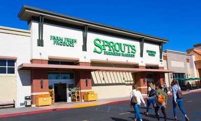 Sprouts Farmers Market at Village at Mira Mesa in San Diego, CA, featuring a white and brick façade with large green signage, wide storefront awning, farm‑fresh produce displays near the entrance, and shoppers walking across the front drive aisle under a vivid blue sky.