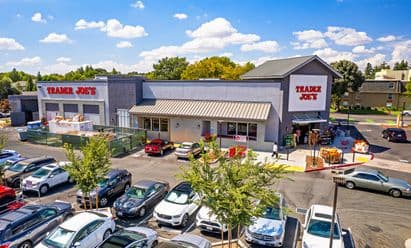 Trader Joe’s at The Davis Collection in Davis, CA, featuring a modern gray gabled façade with large white signage, outdoor seasonal produce displays, bike racks, landscaped walkways, and a wide pedestrian entry area under bright blue skies with scattered clouds.