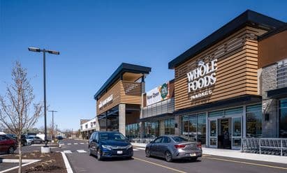Two cars driving in front of Whole Foods Market storefront at Barn Plaza in Doylestown, PA, featuring modern architectural design, active customer traffic, and a newly paved parking area within the retail center.