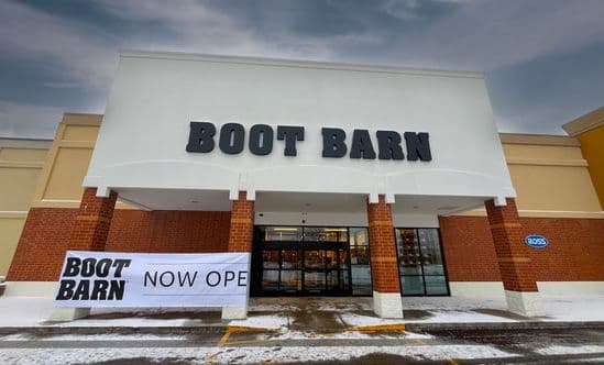 Boot Barn retail storefront with large black signage, brick columns, and glass entrance doors, featuring a ‘Now Open’ banner in front and a snowy parking lot under an overcast sky.