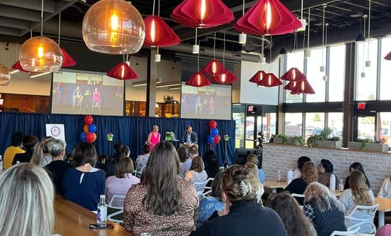 Audience seated at tables watching a live panel discussion in a modern event space with large windows and hanging red pendant lights.