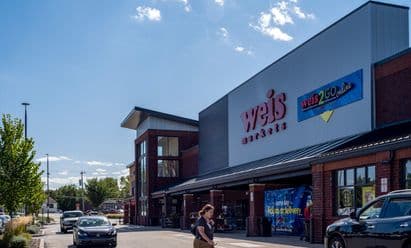 Weis Markets at Plymouth Square in Conshohocken, PA, shown with a contemporary red‑brick and gray metal façade, large glass entryway, covered walkway, prominent ‘weis2GO online’ signage, and vehicles circulating through the front drive aisle.