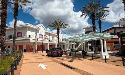 Daytime view of Pointe Orlando in Orlando, FL, featuring a wide pedestrian walkway lined with tall palm trees, a modern glass‑roofed shade structure, landscaped planters, and a two‑story retail and dining building with colorful window graphics and covered storefronts under a partly cloudy sky.