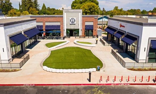 Central courtyard at The Davis Collection in Davis, CA, featuring a large landscaped turf lawn framed by curved concrete seating walls, flanked by two modern retail buildings with white and brick façades, navy awnings, outdoor dining patios, wide pedestrian walkways, and mature trees in the background beneath a bright blue sky.