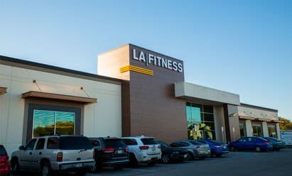LA Fitness at Wynnewood Village in Dallas, TX, featuring a modern façade with wood‑tone vertical paneling, illuminated signage, large glass windows, covered entry walkway, and a busy parking lot with multiple vehicles under a clear blue sky