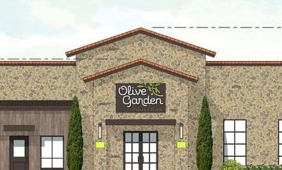 Architectural rendering of an Olive Garden restaurant at Wynnewood Village in Dallas, TX, featuring a stone façade with Mediterranean‑inspired detailing, clay tile roof accents, tall evergreen landscaping, and a prominent branded entryway.