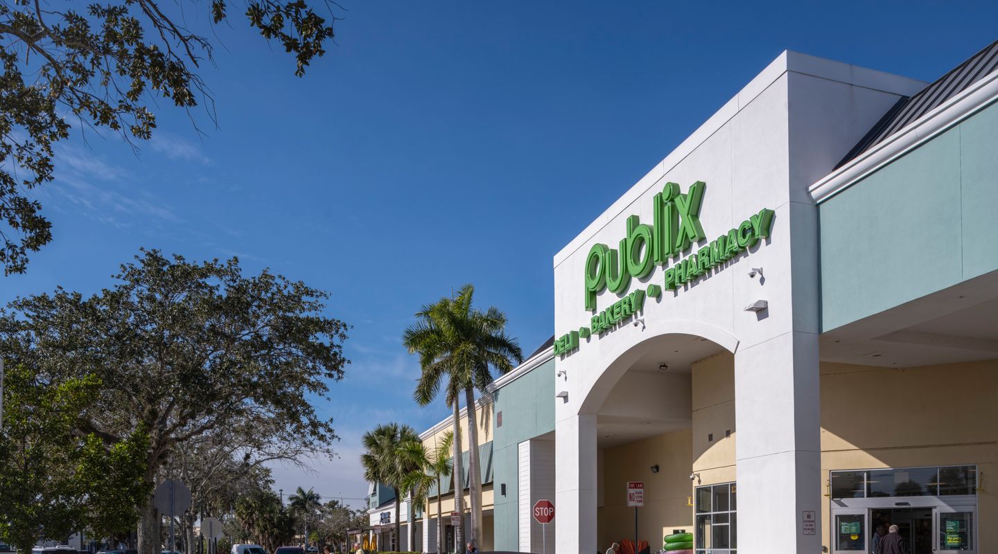 Publix supermarket at Freedom Square in Naples, FL, shown with a clean arched entrance, green signage, landscaped walkways, and shoppers moving through the parking area in front of the store.