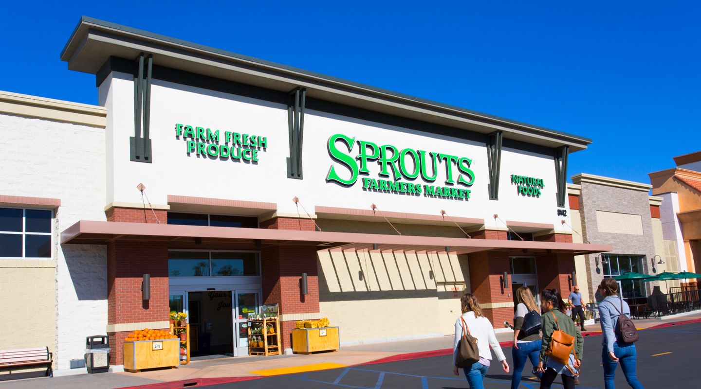 Sprouts Farmers Market at Village at Mira Mesa in San Diego, CA, featuring a white and brick façade with large green signage, wide storefront awning, farm‑fresh produce displays near the entrance, and shoppers walking across the front drive aisle under a vivid blue sky.