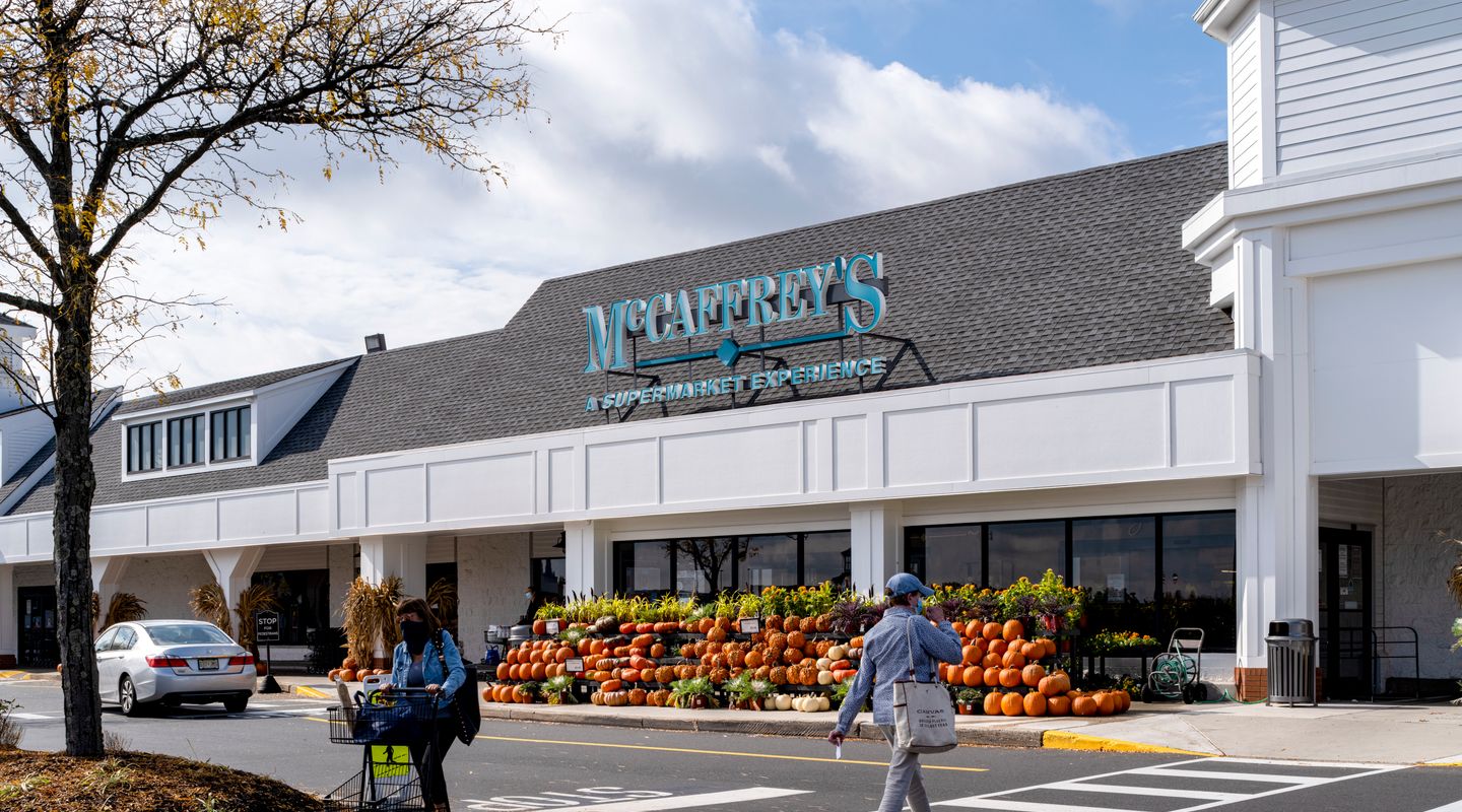 McCaffrey’s Food Market at Village at Newtown in Newtown, PA, shown with classic white clapboard architecture, teal signage, large covered walkway, and seasonal outdoor produce displays including pumpkins arranged along the storefront.