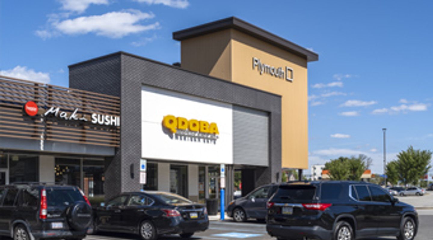 QDOBA Mexican Eats at Plymouth Square in Conshohocken, PA, featuring a modern black brick façade with wood‑accent tower element, adjacent Maki Sushi restaurant, large storefront windows, and a busy parking lot beneath a sunny blue sky
