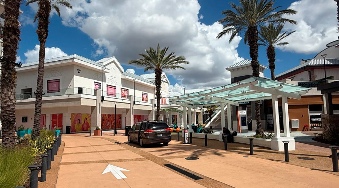 Daytime view of Pointe Orlando in Orlando, FL, featuring a wide pedestrian walkway lined with tall palm trees, a modern glass‑roofed shade structure, landscaped planters, and a two‑story retail and dining building with colorful window graphics and covered storefronts under a partly cloudy sky.