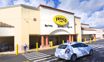 Fresco y Más at Shops at Palm Lakes in Miami, FL, featuring a yellow and cream façade with an arched entryway, bold oval signage, yellow safety bollards, a striped pedestrian crossing, and vehicles entering the front drive aisle beneath bright tropical daylight.