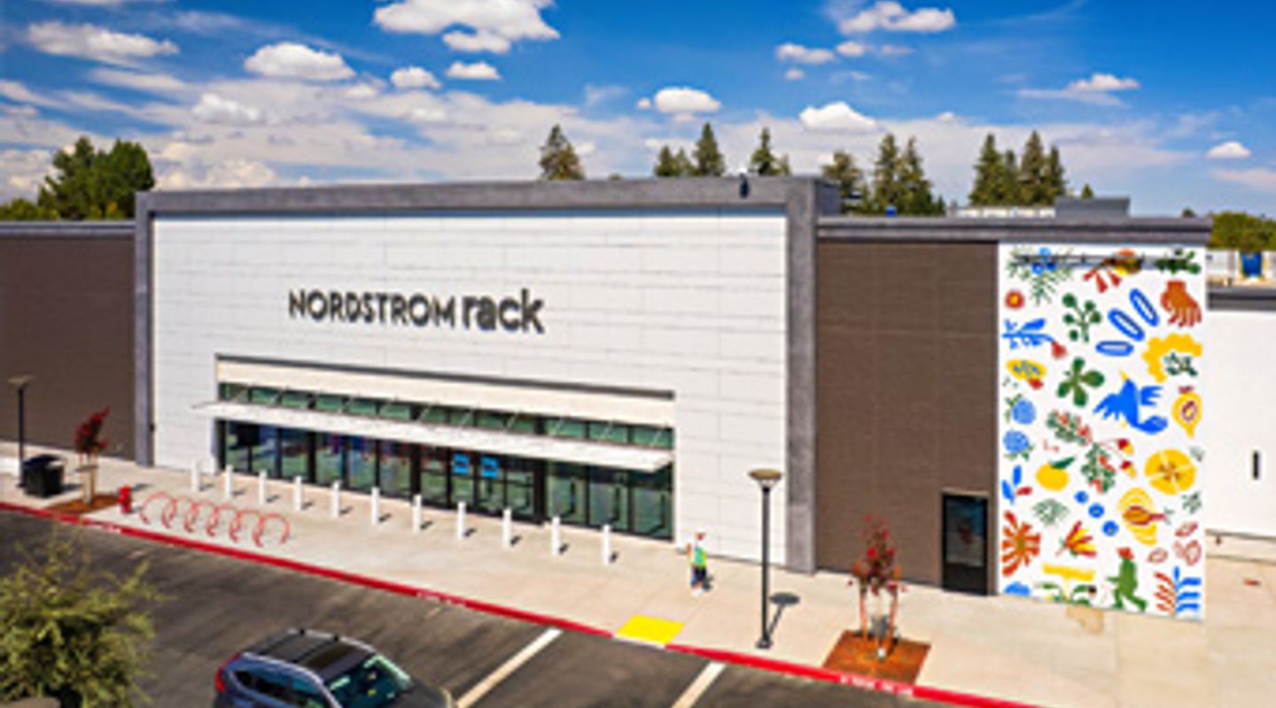 Nordstrom Rack at The Davis Collection in Davis, CA, featuring a sleek modern façade with white architectural panels, large glass storefront windows, bike racks near the entrance, sculptural mural artwork on the adjacent wall, and well‑maintained landscaping under a clear sky.