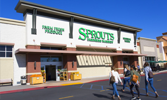 Sprouts Farmers Market at Village at Mira Mesa in San Diego, CA, featuring a white and brick façade with large green signage, wide storefront awning, farm‑fresh produce displays near the entrance, and shoppers walking across the front drive aisle under a vivid blue sky.