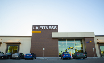 LA Fitness at Wynnewood Village in Dallas, TX, shown with a tall central tower clad in wood‑tone panels, bold illuminated signage, expansive glass entrance framed by light‑colored architectural accents, and parked vehicles in front beneath bright daylight.