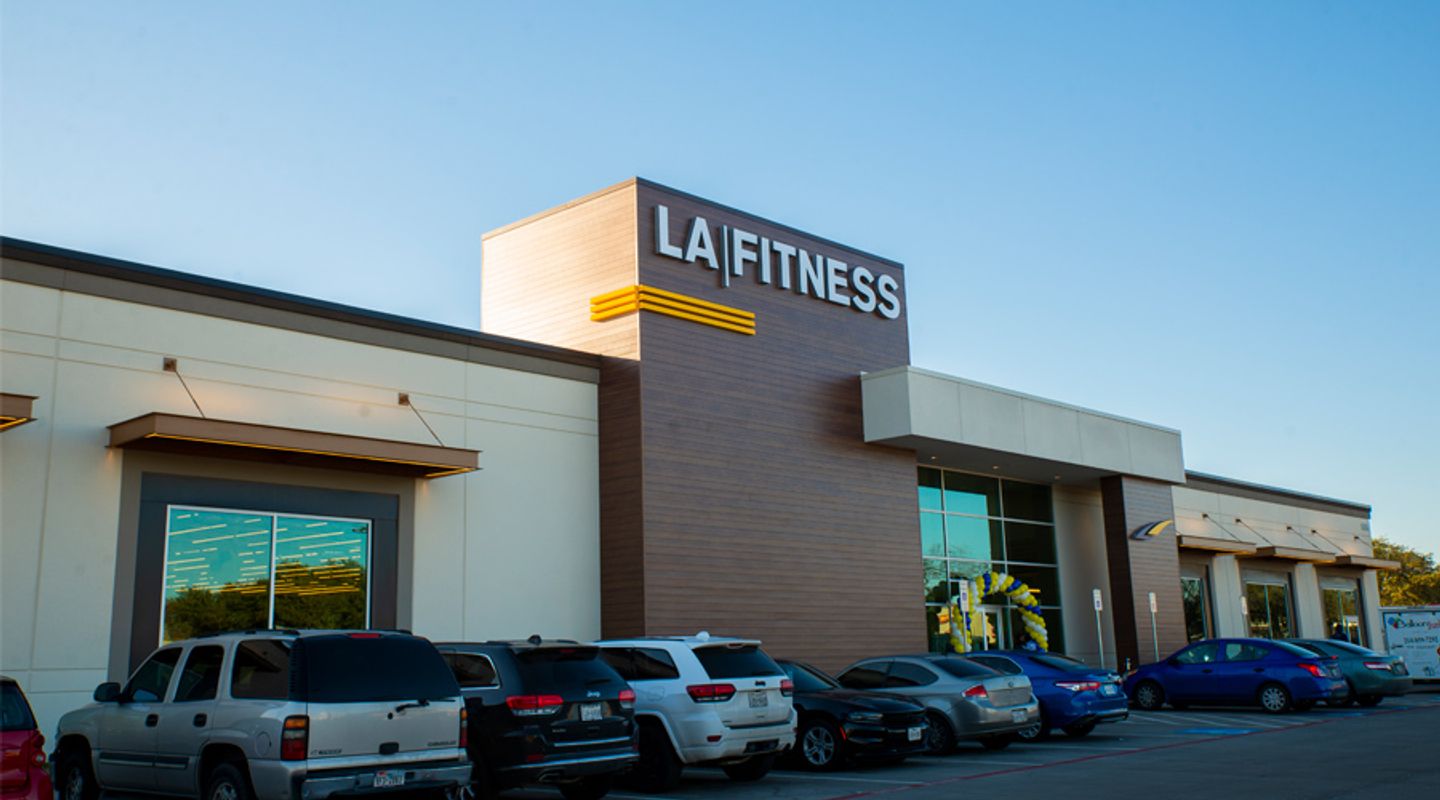 LA Fitness at Wynnewood Village in Dallas, TX, featuring a modern façade with wood‑tone vertical paneling, illuminated signage, large glass windows, covered entry walkway, and a busy parking lot with multiple vehicles under a clear blue sky