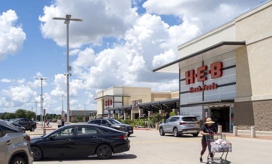 Woman with cart on road in front of busy HEB grocery store.
